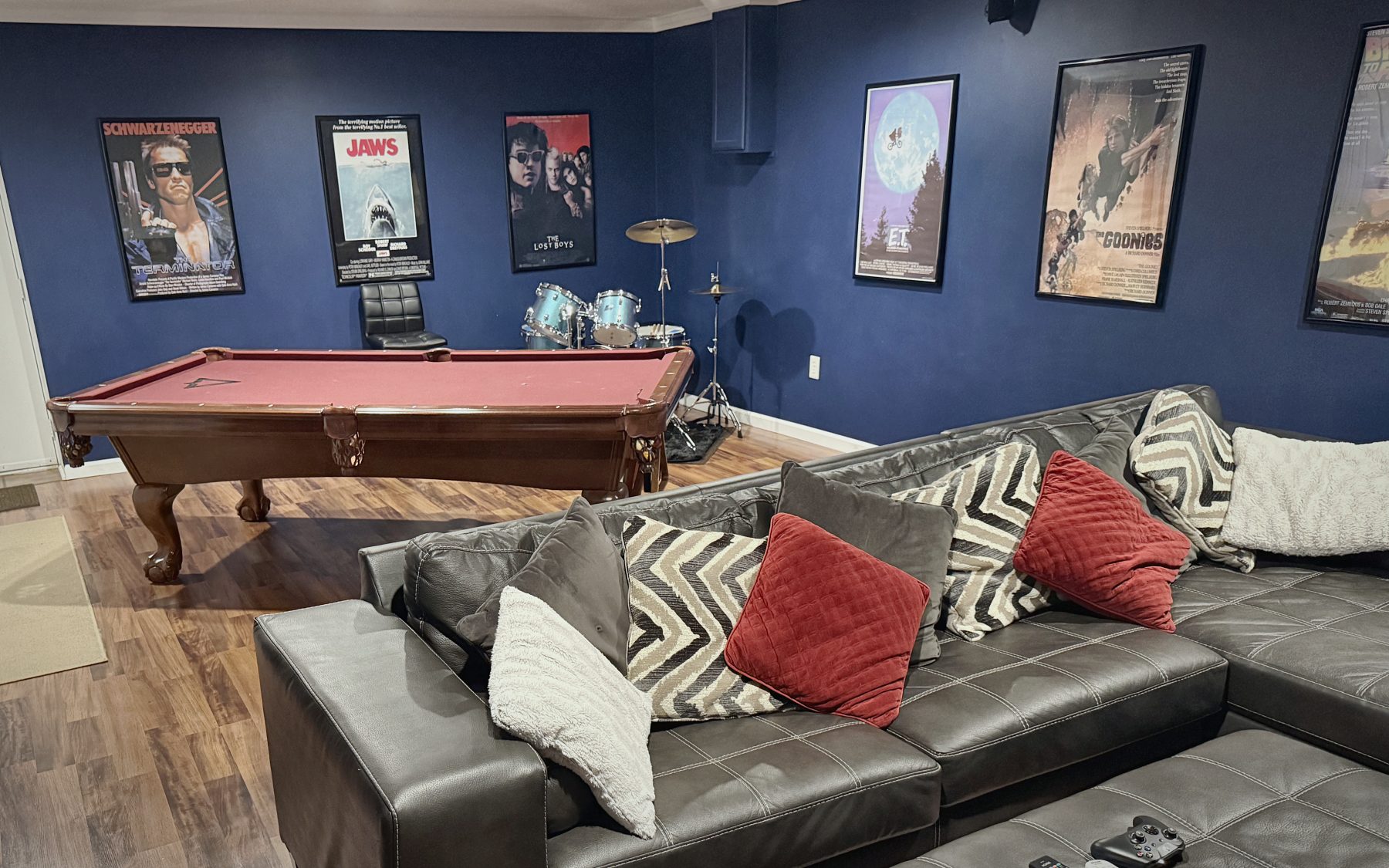 A finished basement games room with a pool table, drum kit, framed film posters and a sectional sofa.