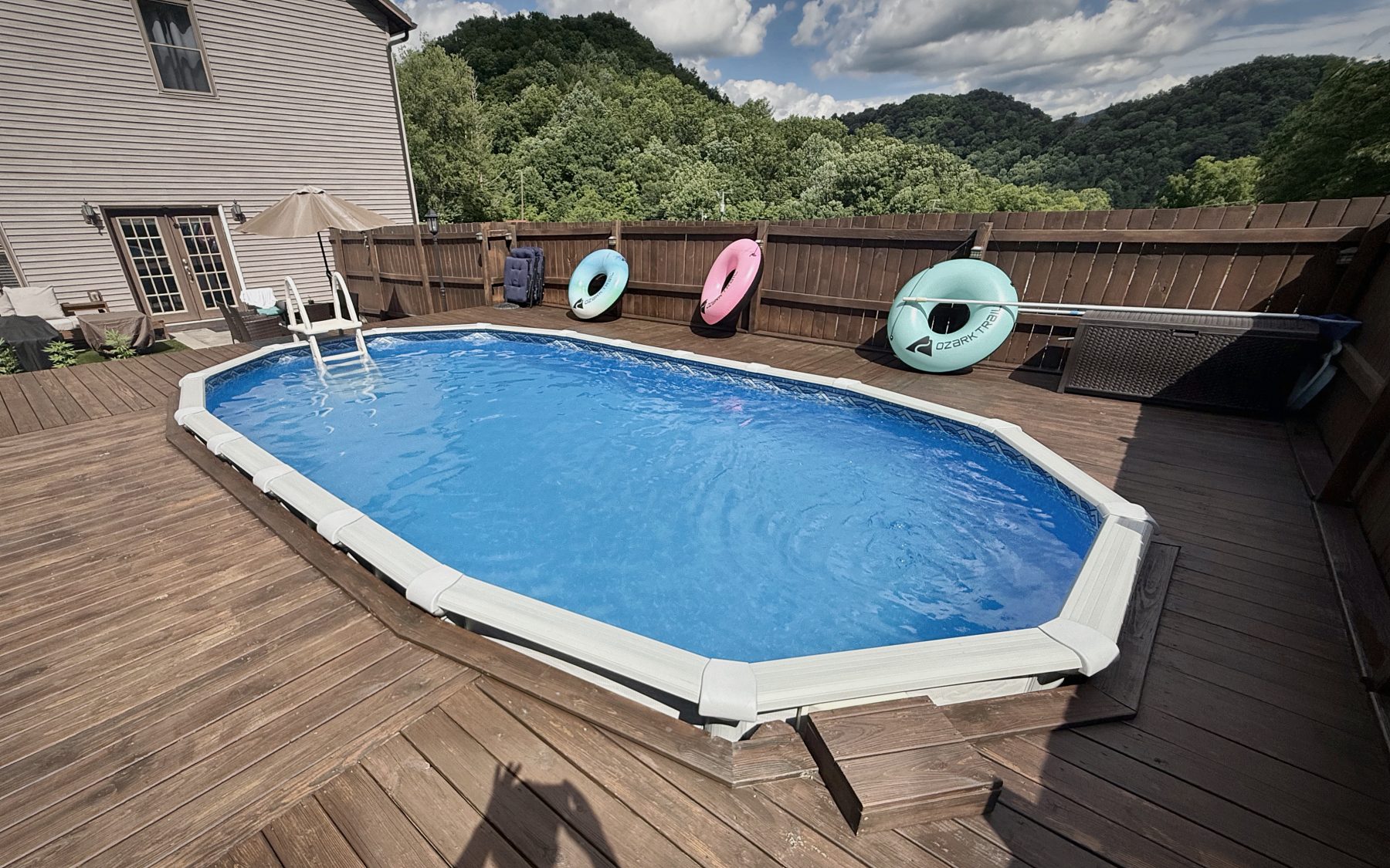 A large timber deck built around an octagonal above-ground pool, the boards cut to follow the pool's angles, with wooded ridgeline behind.