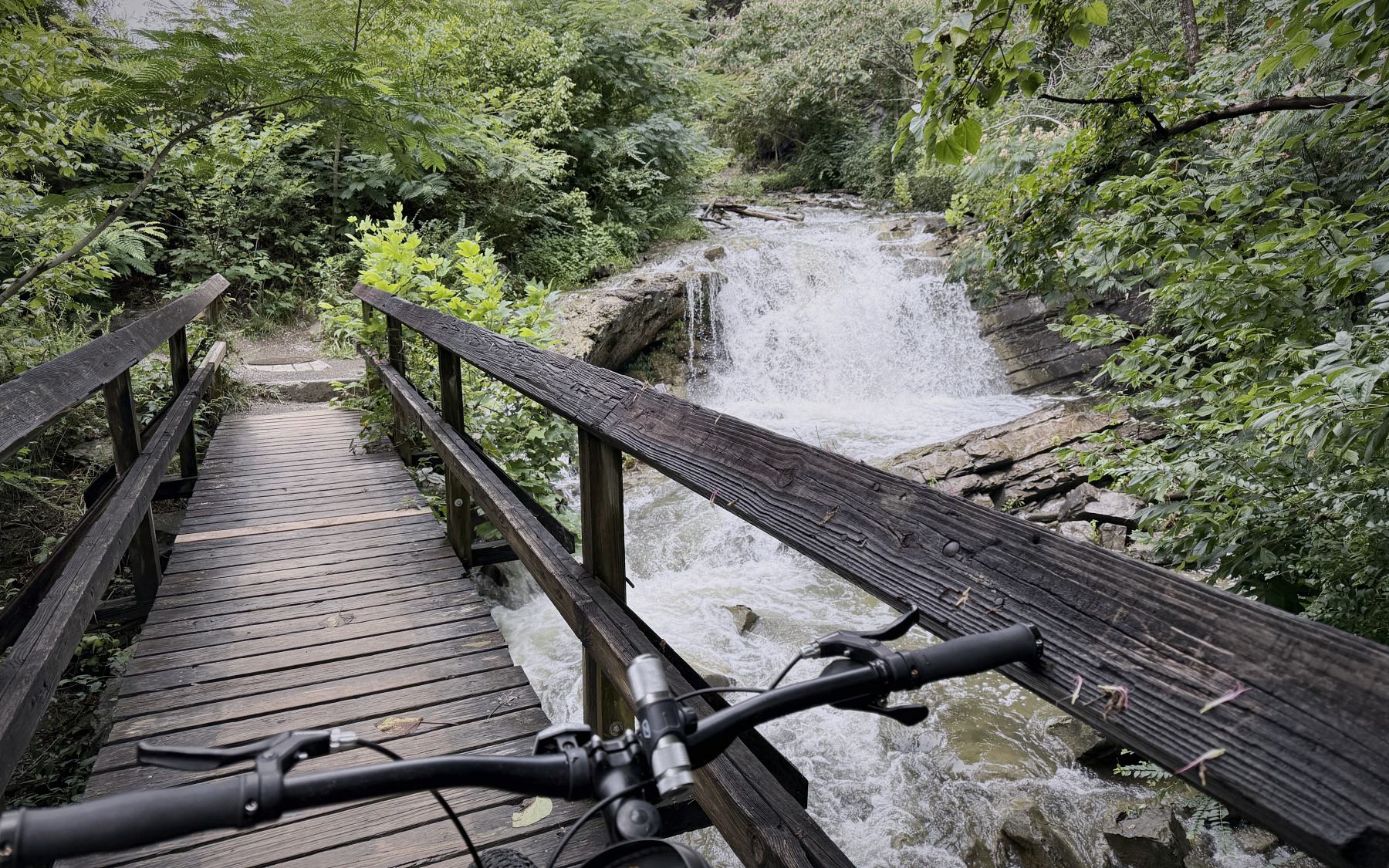 Handlebars of a fat-tire e-bike on a wooden trail bridge, looking out over a waterfall and rapids in dense green woodland.